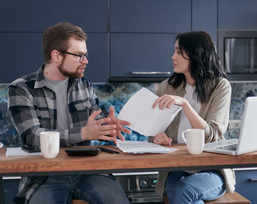 A young couple sits at a table in a bright, modern kitchen, thoughtfully reviewing paperwork, symbolizing the home-buying decision process.